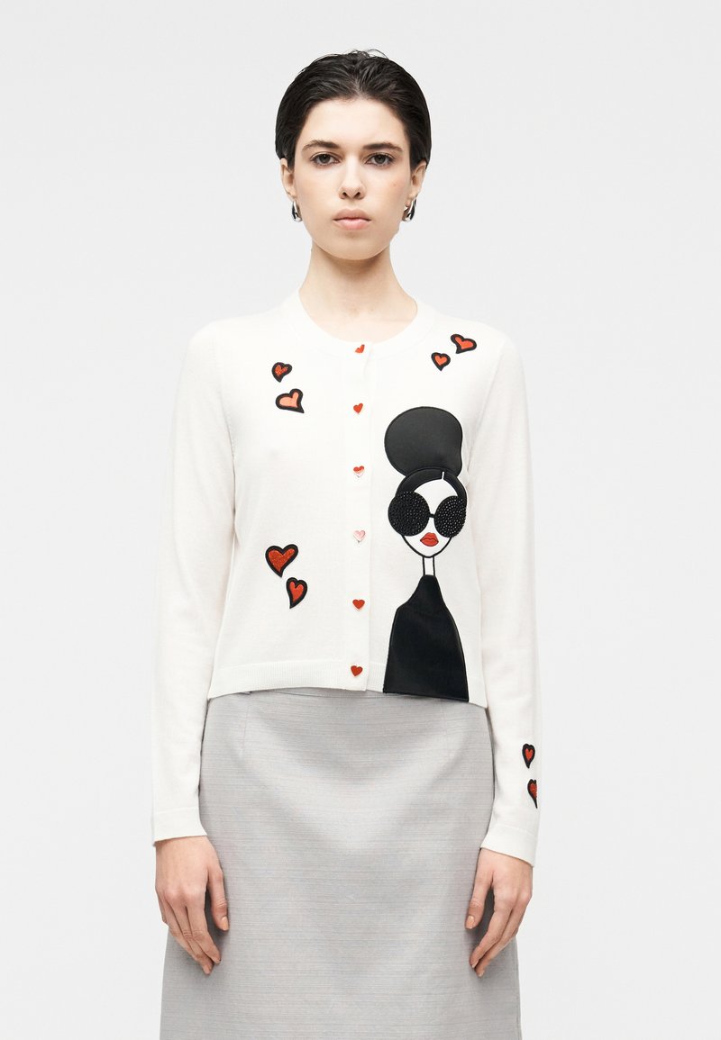 Fehér kardigán piros és fekete szívapplikációkkal, napszemüveges nő sziluettjével. Puha kötött anyag, kerek nyakkivágás és gombos elülső rész.