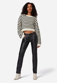 Zwarte leren broek met contrasterende stiksels, gecombineerd met een cropped crèmekleurige trui met zwarte horizontale strepen. Model staat met een zelfverzekerde houding.