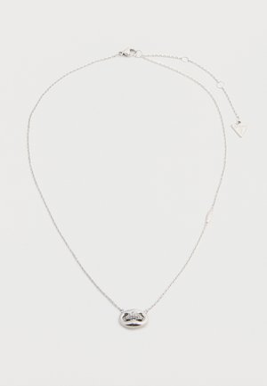 Collier délicat en chaîne argentée avec pendentif ovale orné de cristaux incrustés et une petite étiquette triangulaire de la marque près du fermoir.
