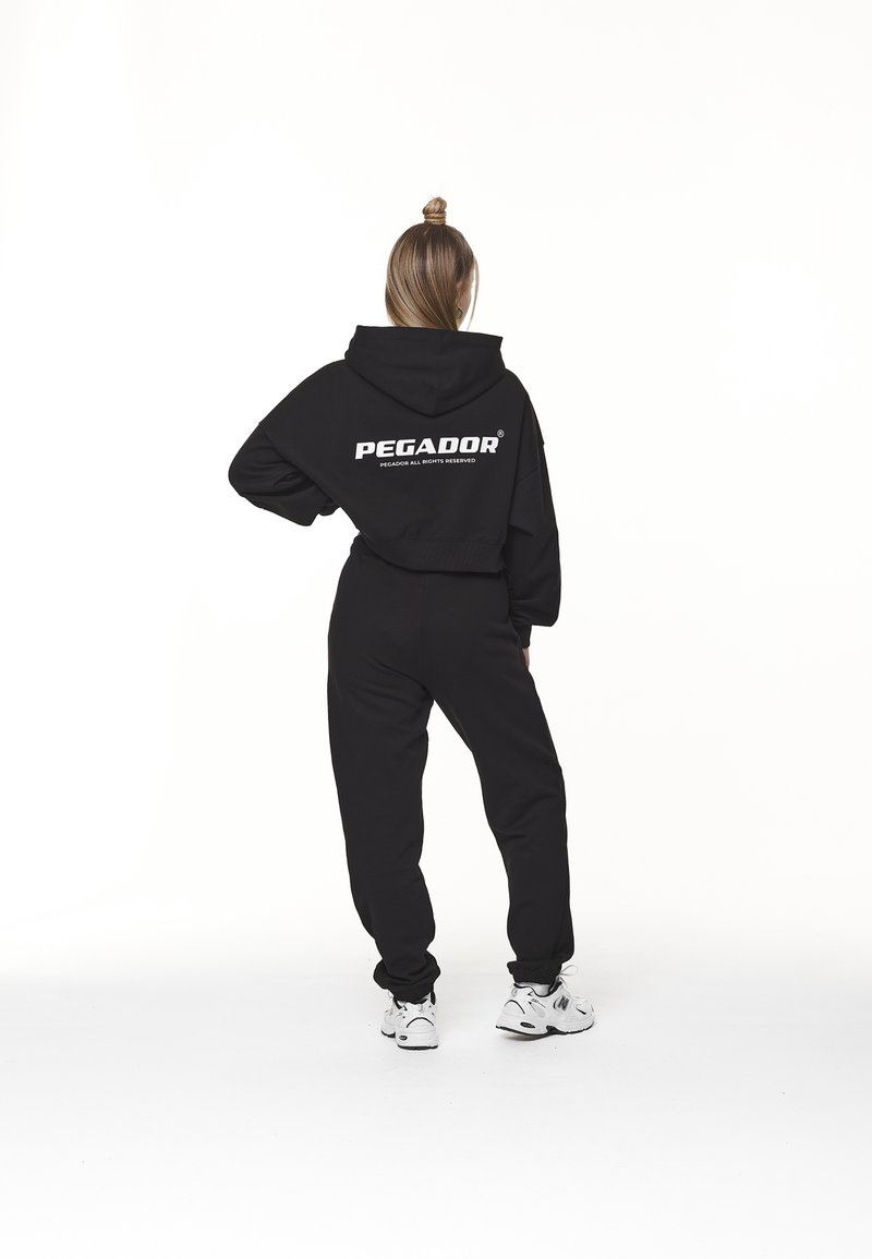 Sudadera corta negra y joggers con el logotipo blanco "PEGADOR" en la parte posterior, combinados con zapatillas blancas, sobre un fondo blanco liso.