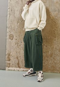 Hellbeige Fleece-Hoodie mit Vordertasche, kombiniert mit grünen Cargohosen mit Seitentaschen und weißen/grauen Nike-Sneakers mit kastanienroten Akzenten.