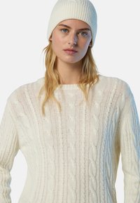 Giovane donna con occhi azzurri che indossa un berretto di lana bianco e un maglione a trecce color crema, rivolta verso l'avanti su uno sfondo neutro.