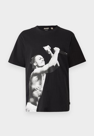 Svart oversized t-shirt med en stor svartvit grafisk bild av en barbröstad figur som håller i en mikrofon. Tillverkad av bomullstyg.