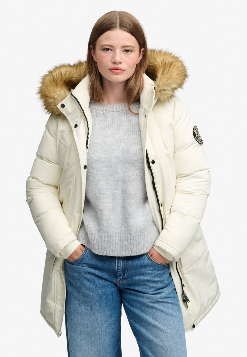 Woman wearing a cream puffer jacket with fur-lined hood, light gray sweater, and blue jeans, hands in pockets, standing against plain background.