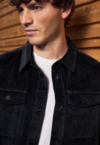 Mladý muž s světlou pletí a kudrnatými hnědými vlasy, oblečený v černé manšestrové bundě přes bílé tričko, stojící proti dřevěné zdi.