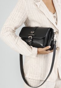 Svart läder crossbody-väska med klaffstängning, silverfärgad hårdvara, justerbar rem och präglad logotyp på framsidan.