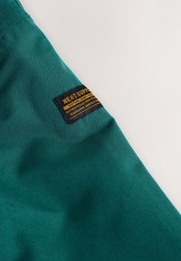 Tmavozelená látka s prišitou obdĺžnikovou čierno-žltou štítkou s nápisom "NEXT SUPPLY Established 1982 Authentic Crafted."