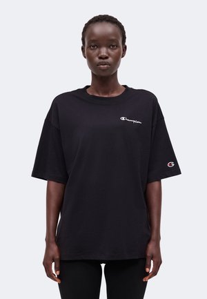 Ung kvinde iført en sort oversized Champion t-shirt med logoer på bryst og ærme, stående mod en ensfarvet lys baggrund.
