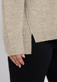 Maglione beige lavorato a maglia con polsini a coste e spacchi laterali, mostrato mentre è tenuto da una mano. La texture appare morbida e calda su un tessuto nero.