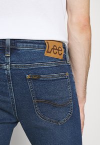 Blue denim jeans with a brown leather patch labeled "Lee" on the waistband. Features yellow stitching and a classic five-pocket design.