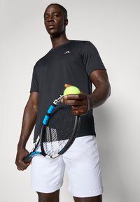 Sort atletisk T-shirt med et lille logo, hvide shorts, holder en tennisracket med et blåt og sort håndtag, og en grøn tennisbold.