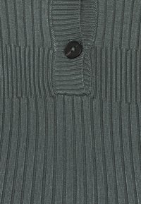 Pull gris côtelé avec un bouton noir au niveau de l'encolure, présentant un design texturé mettant en valeur des lignes verticales.