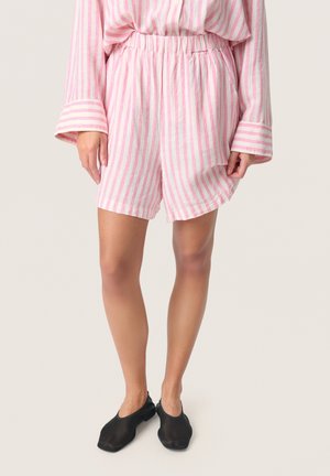 Persoon draagt losse roze en witte verticale gestreepte korte broek en bijpassend langärmelig shirt met zwarte instapschoenen, staand op een lichte achtergrond.