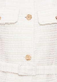 Σακάκι από ύφασμα με κρεμ υφή, διακοσμημένο με χρυσά κουμπιά με ανάγλυφο μοτίβο, τσέπες με καπάκι και ζώνη από ίδιο ύφασμα στη μέση.