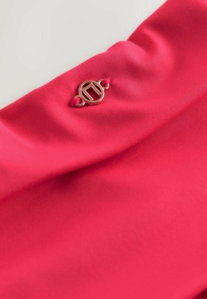 Roosa kangas, mis näitab sujuvat tekstuuri ja mille külge on ühendatud kuldse tooniga logo detail punase nööri abil, esitledes volditud disaini puhast väljanägemist silmas pidades.