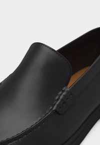 Mocassin en cuir noir à enfiler avec dessus lisse et coutures visibles le long des bords, montrant une partie de la doublure intérieure couleur tan.