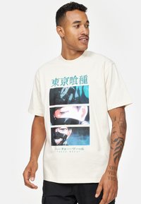 Beige bomulds t-shirt med et grafisk print af tre anime-inspirerede ansigtstræk i teal og sort. Kortærmet, rund halsudskæring og afslappet pasform.