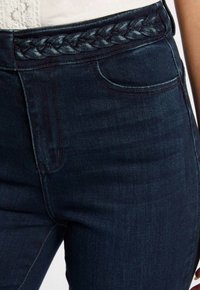 Jean en denim foncé avec une taille tressée ; design à cinq poches avec des détails de couture subtils ; texture lisse et silhouette ajustée.