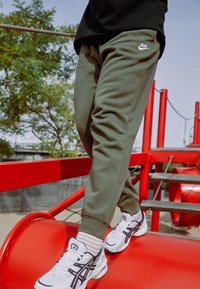 Persoon in groene Nike-sweatpants en witte sneakers die in balans staat op een rode speelstructuur onder een blauwe lucht.