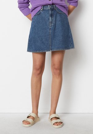 Personne portant une jupe trapèze en denim bleu, un pull en tricot violet et des sandales beiges à double bride, debout devant un fond blanc.