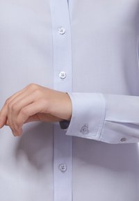 Hellblaues Button-up-Hemd aus Stoff, mit glatter Textur, Manschetten mit Knopfdetails und taillierter Passform.