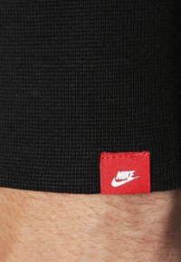 Černá pletená látka s texturovaným vzorem, která má malou červenou tkanou etiketu s bílým logem Nike.