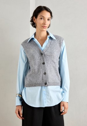Gilet sans manches en laine grise avec un col en V et cinq boutons foncés, porté sur une chemise à manches longues bleu clair. La chemise a une coupe ample.