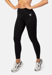 Čierne legíny s vysokým pásom a hladkým povrchom, s malým bielym logom na ľavom boku. Skombinované s robustnými bielymi a čiernymi teniskami.