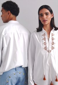 Deux modèles portent des chemises blanches. L'une présente des broderies marron et des franges. L'autre est unie avec un col classique. Des jeans en denim sont visibles.