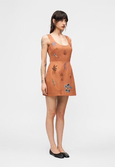 Γυναίκα με σκούρα μαλλιά που φοράει καφέ μίνι φόρεμα χωρίς μανίκια με πολύχρωμα κεντημένα αφηρημένα σχέδια και μαύρα επίπεδα παπούτσια, στέκεται μπροστά σε απλό φόντο.