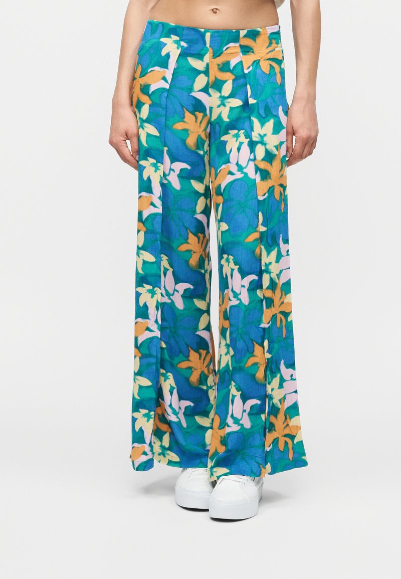 Άτομο που φοράει παντελόνια με ευρεία μπατζάκια, με πράσινο, μπλε, κίτρινο και ρόζ λουλουδάτο μοτίβο, σε συνδυασμό με λευκά παπούτσια πλατφόρμας σε λευκό φόντο.