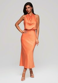 Robe sans manches à col halter en tissu orange doux et texturé, avec un corsage ajusté et une jupe fluide arrivant aux chevilles. Talons nus à brides.