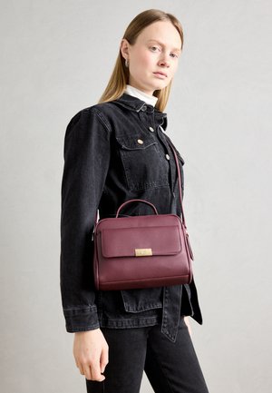 Sac à main en cuir bourgogne avec fermoir en or, forme structurée, anse supérieure et bandoulière réglable. Le mannequin porte une veste en jean.