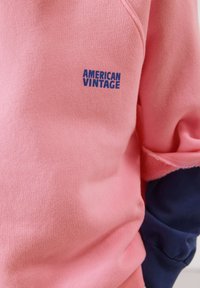 Gros plan sur un sweat-shirt rose avec le texte bleu « AMERICAN VINTAGE » et un bord-côte bleu marine visible sur le bras droit.