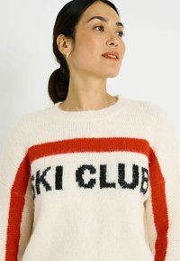 Suéter crema con una franja roja y "SKI CLUB" tejido en negro. Textura suave, cuello redondo y ajuste relajado con hombros caídos.
