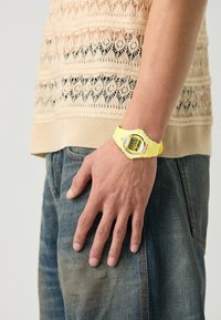 Digital watch with a bright yellow silicone strap and a digital display, worn on a hand against a light-colored knitted top and denim jeans.