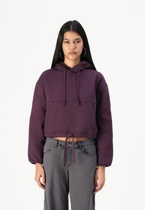 Violets krāsas apgrieztais hoody ar aukliņu apakšmalā, ar priekšējo kabatu un izšūtu logo uz kreisā piedurkne. Kokvilnas maisījuma audums.