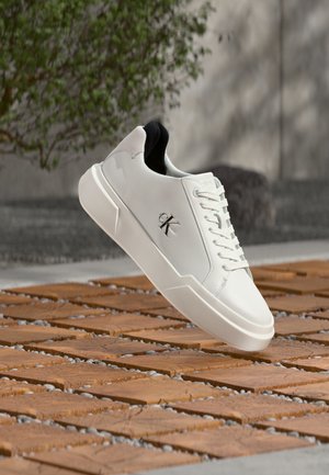 Baskets blanches en cuir Calvin Klein avec une semelle épaisse flottant au-dessus de carreaux en bois et de petits cailloux en plein air près d'une plante verte et d'un mur en béton.