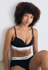 Black bralette with adjustable straps and a white Calvin Klein logo band, paired with matching high-waisted black briefs. Smooth texture.