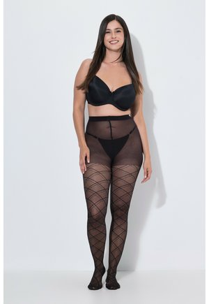 Frau, die steht und lächelt, trägt schwarzen BH und gemusterte schwarze transparente Strumpfhosen vor einem einfarbigen hellen Hintergrund.