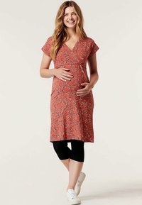 Vestido de maternidade em coral com estampado de folhas, decote em V, mangas curtas, corpete justo e parte inferior ampla. Combinado com leggings pretas e sapatilhas brancas.