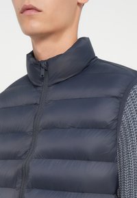 Nahaufnahme einer Person, die eine marineblaue gesteppte Weste über einem grauen Strickpullover trägt, wobei der Fokus auf dem Reißverschluss der Weste und dem hohen Kragen liegt.