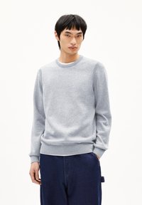 Hellgrauer Pullover aus weichem Strickstoff, mit Rundhalsausschnitt, gerippten Bündchen und Saum sowie glatter Textur. Getragen mit blauen Hosen.