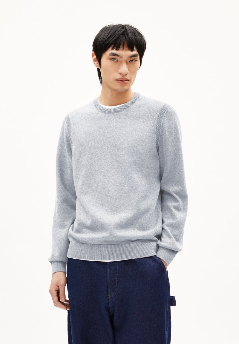 Hellgrauer Pullover aus weichem Strickstoff, mit Rundhalsausschnitt, gerippten Bündchen und Saum sowie glatter Textur. Getragen mit blauen Hosen.