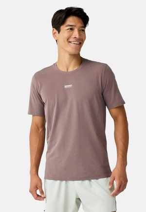 Uomo sorridente che indossa una maglietta malva strutturata con un piccolo logo bianco "BROOKS" e pantaloncini chiari su uno sfondo semplice.
