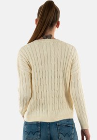 Suéter de punto trenzado en color crema, con puños y dobladillo acanalados. Patrón texturizado con puntos alargados, llevado sobre jeans azules.