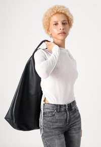 Sac fourre-tout en cuir noir avec une surface texturée, grande taille et design minimaliste, porté sur l'épaule. Le modèle porte un haut léger et ajusté.