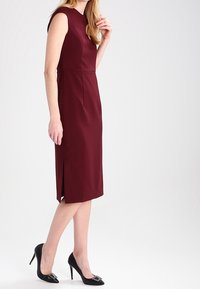 Bordeaux mouwloze jurk met een aansluitende silhouet, knielang, met een split aan de zijkant en een gladde stof. Gecombineerd met zwarte hoge hakken met versieringen.