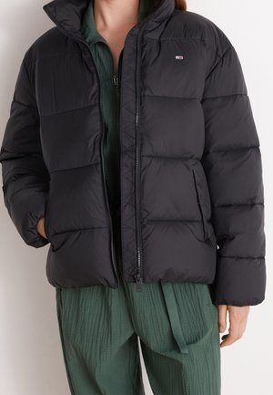 Person iført en sort dunjakke over et grønt tekstureret zip-up outfit med hænderne delvist i jakkelommerne.