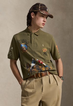 Polo vert olive avec des motifs en relief, des motifs de cheval et cavalier, et des logos brodés sur les manches. Associé à un pantalon beige et une ceinture marron.